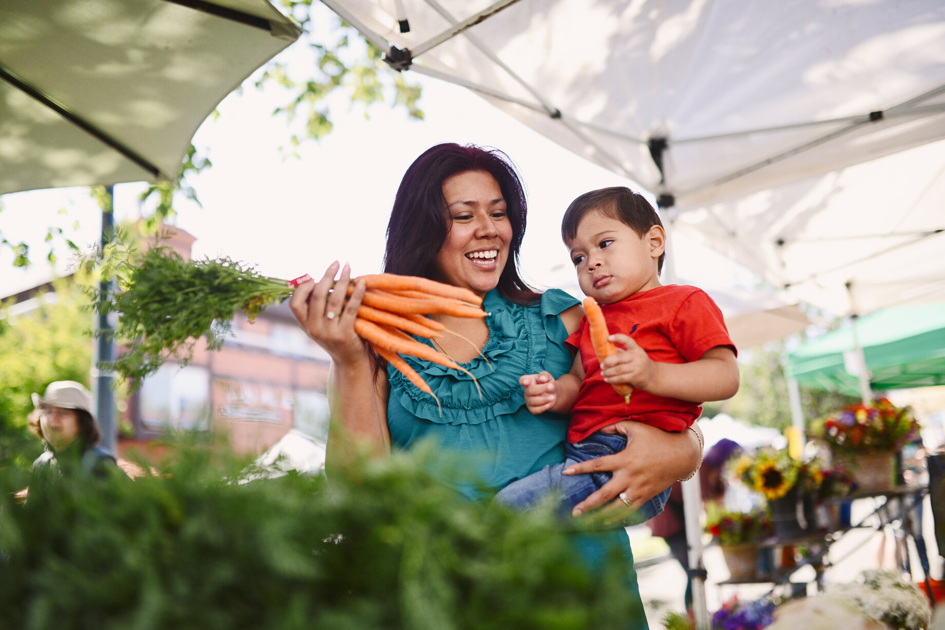 Mother showing toddler son a bunch of carrots at a farmers market